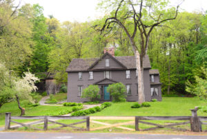 Old home in Concord, MA with dark brown siding and a blue front door with beautiful green grass and trees around the property