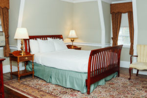 Historic Queen bed guestroom dated back to 1716 in Concord, MA with original pine plank floors, wainscoting, queen bed with white comforter with wooden bed frame, wooden bed side table and white chair