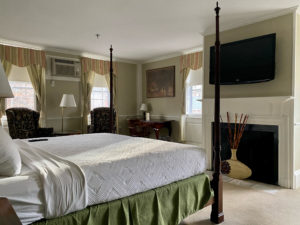 King bed guest room with four post wooden bed frames, white comforter with green bed skirt, flatscreen tv hanging on the wall above the fireplace, two dark floral pattern chairs and deck in the corner in Concord, MA hotel