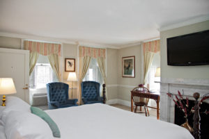 Accessible Suite king room in Concord, MA hotel with four wooden post bedframe and white comforter, two blue oversized chairs, desk area and flat screen tv hanging above the fireplace