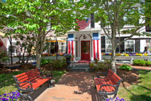 Brick entranceway into the Inn with 2 red benches, planted flowers, and bloomed trees with 2 American flags hanging on each side of the door and an American flag hanging above the front door in Concord, MA