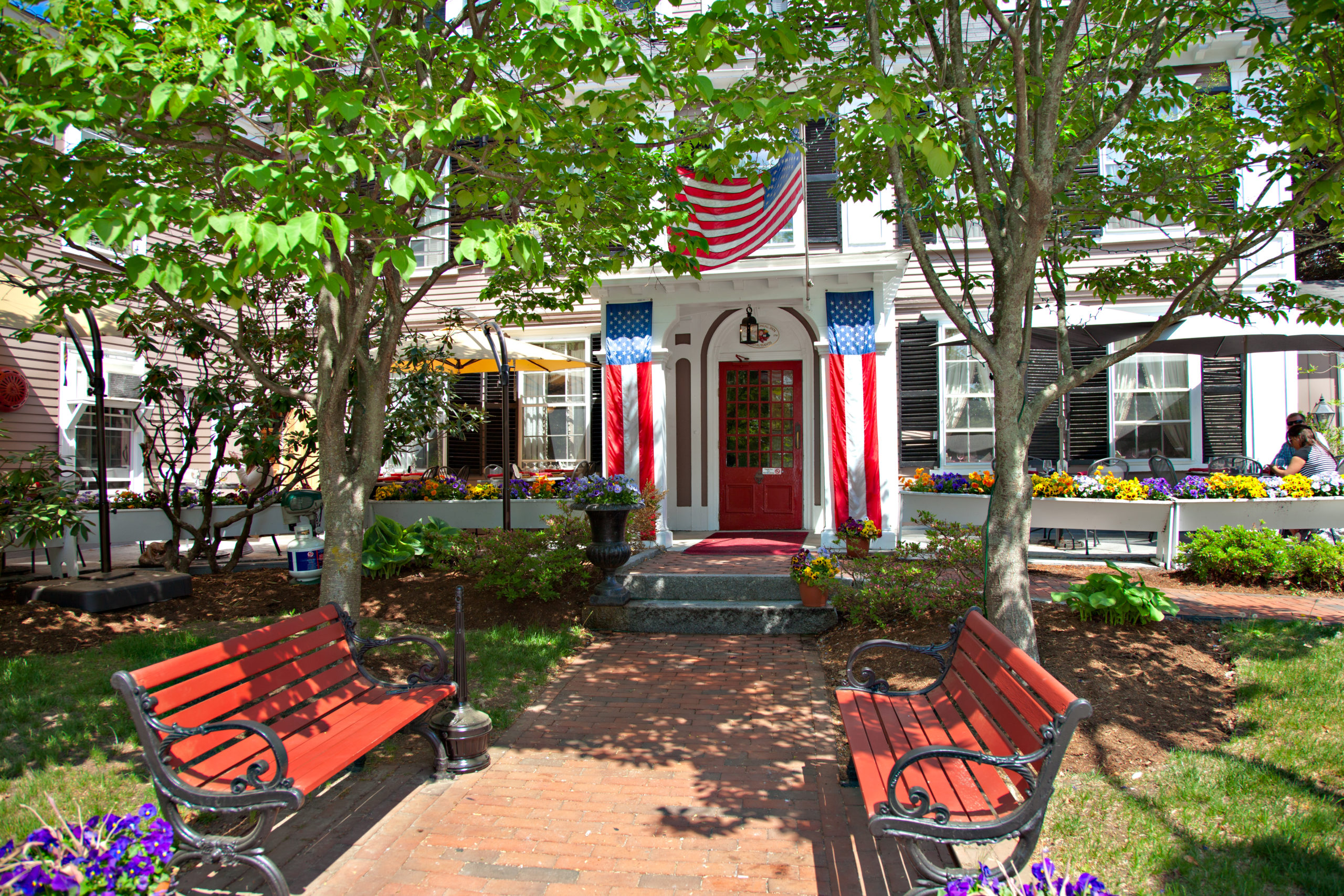 Brick entranceway into the Inn with 2 red benches, planted flowers, and bloomed trees with 2 American flags hanging on each side of the door and an American flag hanging above the front door in Concord, MA